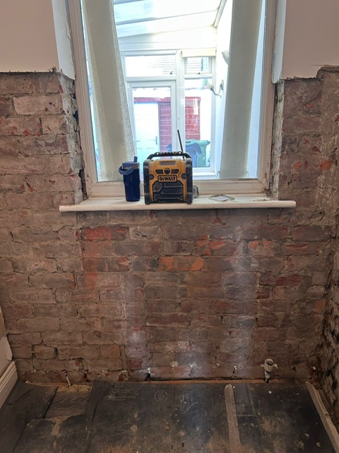 A wall hacked back to brick around a window with a damp meter on the sill