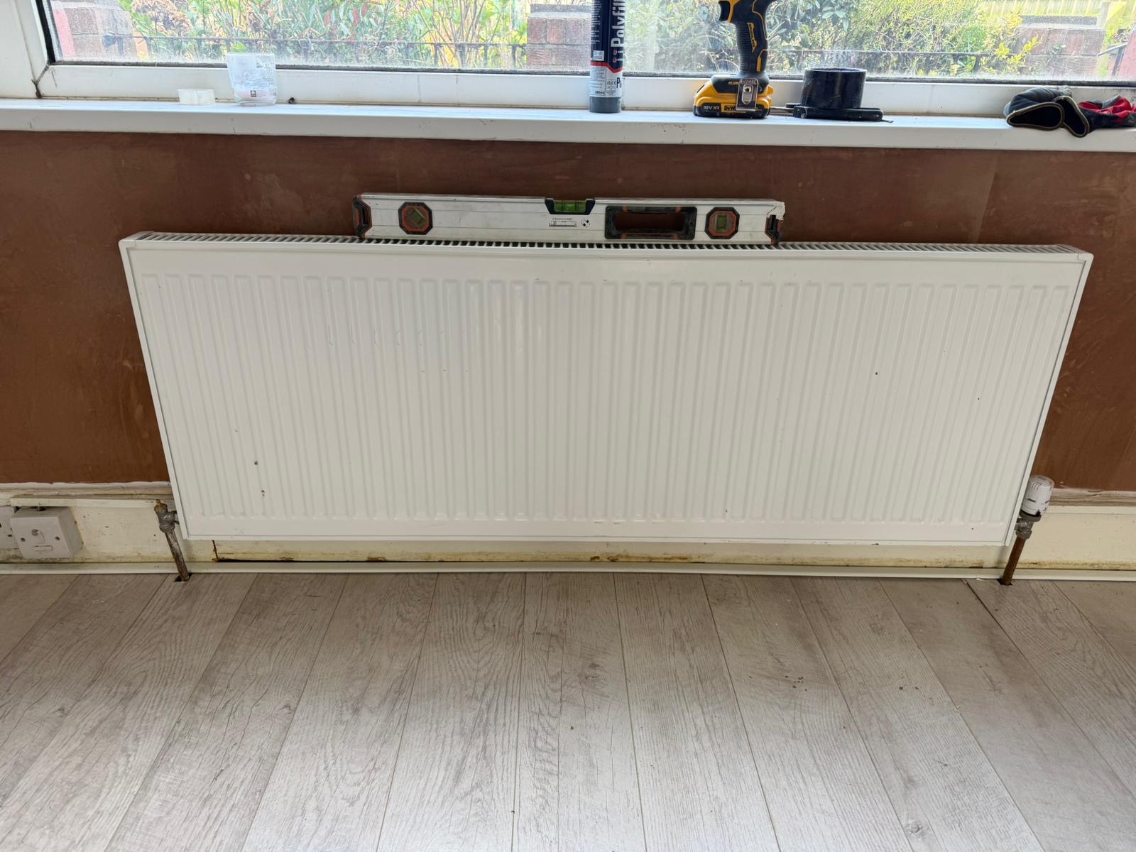 A wall below a window stripped back ready for damp proofing, with the radiator in place and the floor protected